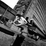 A man with dreadlocks is captured mid-air during a parkour jump between two concrete structures in an urban setting. He is extending his arms forward, wearing a white T-shirt and track pants. A tall building and other people are visible in the background. Black and white image.