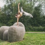 A man performs an athletic, one-handed handstand on large stone blocks in a grassy park area surrounded by trees and bushes. He is shirtless, wearing light pants and sneakers, his body creating a dynamic pose against the green backdrop.