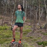 A person with long dreadlocks, wearing a green shirt, black shorts, and bright orange running shoes stands on a tree stump in a forest. They look thoughtfully to the side while surrounded by bare trees and moss-covered ground.