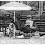 A black-and-white photo of a street musician sitting on a blanket in front of a van, playing a pan flute. A striped umbrella provides shade, and a wheelchair is positioned nearby. The musician wears a striped hat and appears deeply focused on playing.