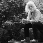 A man with long white hair and a beard sits thoughtfully on a wooden bench next to a bicycle. He is wearing a button-up shirt, pants, and casual shoes. The background is filled with dense foliage, and the scene is in black and white.