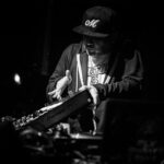 A DJ, wearing a baseball cap and hoodie, intensely focuses on mixing tracks on his Pioneer DJ controller in a dimly-lit scene. He has one hand on the controller and the other hand gesturing above it, creating an engaging atmosphere.