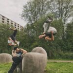 Three people perform parkour stunts in an outdoor setting with large stone structures. One person flips backward in midair, another crouches on top of a stone, and the third flips forward. Trees and a building are visible in the background.