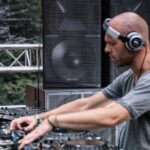 A bald DJ wearing a gray T-shirt and headphones is focused on mixing music on a turntable at an outdoor event. Large speakers and various audio equipment surround him. The background is blurred but shows trees and metal stage structure.