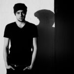 A black and white photo of a man standing against a wall, casting a shadow. He is wearing a dark T-shirt, beanie, and jeans, with his hands in his pockets. The lighting creates strong contrast, emphasizing the shadow on the wall.