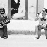 A man wearing a T-shirt, shorts, and sunglasses sits on a bench outside, drinking from a bottle. Next to him on the bench is a bronze statue of an elderly man with a beard holding a hat in his lap. In the background, there's an advertisement featuring a woman's image.