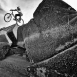 A cyclist wearing a helmet performs a daring stunt, riding on one wheel over massive, rugged boulders by the beach. The sun's rays penetrate from the top right, illuminating the scene with a dramatic flair, emphasizing the cyclist's agility and balance.