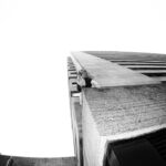 A person is seen hanging off the edge of a tall building, with one hand clutching the corner and wearing a cap, viewed from below in dramatic perspective. The photo is shot in black and white, enhancing the stark contrast and height.