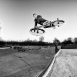 Black and white image of a person performing an aerial trick on a BMX bike in a skatepark. The rider is high above the ramp, holding the handlebars with both hands while the bike is parallel to the ground. Graffiti decorates the walls of the skatepark.