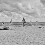 Black-and-white photo of a beach scene. Several people are scattered across the sandy shore, some sitting on towels or sunbathing. A few are walking along the water's edge, and a person carrying bags walks through the scene. The sky is cloudy.