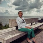 A person with short hair, wearing a white patterned blouse, turquoise pants, and dark shoes, sits on a wooden bench by the water. The sky is filled with dramatic clouds, creating a contrasting background. The person is looking confidently towards the camera.