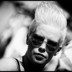 Black and white photo of a person with short, spiky hair wearing heart-shaped sunglasses. The person’s expression is neutral and the background is blurred, suggesting depth. The image has a high contrast, emphasizing shadows and highlights.