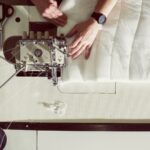 A person operates a sewing machine, guiding white fabric under the needle. Spools of white thread are mounted on the left side of the machine. The individual's hands are actively working with the fabric while wearing a watch on the left wrist. Scissors and measuring tape lie nearby.