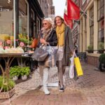 Two women, one older and one younger, walk arm in arm down a charming, narrow street with shopping bags in hand. They're both smiling and appearing joyful. The street is lined with brick buildings and vibrant storefronts, including a flower shop.