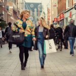 A woman and a young girl, carrying shopping bags, walk hand-in-hand down a busy pedestrian street. They are both smiling and appear to be enjoying their day. The street is lined with various shops, and other people walk or bike in the background.