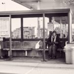 A black and white photo of a person sitting alone on a bench inside a bus stop shelter. The person is bundled in warm clothing, and there are bags beside them. The backdrop includes a gas station and urban buildings. There are signs and a trash can nearby.