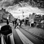 A black and white photo of a busy street scene. People are walking along the sidewalk near a row of parked bicycles. European-style buildings line the street, and the sky above is filled with dramatic clouds. A reflective surface captures a portion of a car in the foreground.