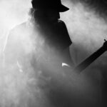 A black and white photo of a person with long hair and a beard, wearing a cap, playing an electric guitar. The image is shrouded in smoke or fog, and the figure is mostly in shadow, giving a mysterious and atmospheric effect.