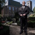 An elderly man in a dark suit stands solemnly in a churchyard, holding a black hat. He is positioned in front of an old brick church with a tall steeple, surrounded by gravestones, flowers, and trees. The ambiance is somber and reflective.