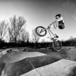 A person wearing glasses performs a high jump on a BMX bike at a skatepark. The black-and-white photograph captures them mid-air above a graffiti-covered ramp, with bare trees and a clear sky in the background.