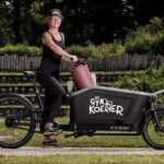 A person in athletic wear stands beside a cargo bike with a large front box. The box displays the text "De Gym Koerier." The person, wearing a cap, holds a yoga mat. The background features greenery and a wooden fence.