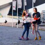 Two young women stand outside a modern building. One is holding a skateboard and the other is on a skateboard. They are both looking at a map and smiling, wearing casual outfits. Several flags can be seen in the background.