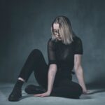 A person with shoulder-length blonde hair, wearing black-rimmed glasses, sits on the floor against a dark grey background. They are dressed in black clothing and black boots. Their arms, which have visible tattoos, rest on the floor and their knee, gazing downward.