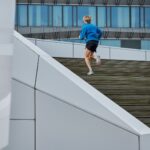 A person wearing a blue jacket and black shorts is seen from behind, running up a long set of wooden stairs outdoors. The background features modern, angular white architecture and a glass building with vertical blue windows.