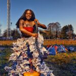 A person with long hair stands outdoors on a grassy field, holding a rope. They wear a dress made of flowing, crumpled paper and an orange-red top. Behind them are scattered electronic devices, and a tall communication tower is visible in the background. The sky is clear.