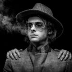 A black-and-white image of a person wearing a woven hat, aviator sunglasses, and a coat. They stare into the distance while a pair of hands rests on their shoulders, with smoke drifting to the left side of the image. The background is completely black.