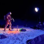 A person wearing a coat, hat, and gloves stands on a beach at night, holding a light source. They are surrounded by several pieces of lighting equipment, some providing blue and red light, and a large spotlight on a stand is illuminating the scene.