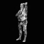 Black and white image of a heavily tattooed individual standing against a dark background. They have a long, full beard, glasses, and tattoos covering their face, arms, torso, and legs. They wear camouflage shorts and rest one arm on their head while the other arm is on their chest.