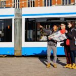 Three people stand near a GVB tram. One person is pointing in a direction while holding a map, another has a camera hanging around their neck, and the third person has a blue backpack. They appear to be tourists, possibly figuring out directions.