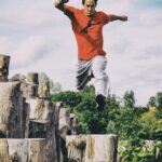 A person wearing a red shirt and gray pants is captured in mid-air, leaping from one wooden stump to another in an outdoor setting surrounded by greenery and a cloudy sky.