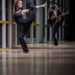 A person is performing a standing split while balancing on one leg in a dance studio. They are dressed in a dark, patterned outfit. The pose is mirrored on the glass wall behind them, showcasing both their flexibility and strength.