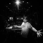 A DJ wearing a hat and a T-shirt stands behind his equipment, arms outstretched towards the camera, performing in a dimly lit venue. The crowd is visible in the background, and a spotlight shines brightly above his head, casting dramatic lighting.