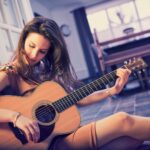 A young woman is sitting on the floor, leaning against a doorway, playing an acoustic guitar. She has long hair and is wearing a short dress. The room is softly lit, and a window with sheer curtains is visible in the background.