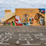 A vibrant mural covers an orange building with various colorful artwork, including abstract faces and animals. "Feliz Natal" is painted on the ground before the building. The sky is partly cloudy, and there's a person in the bottom left corner near the mural.