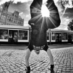 A person performs a handstand on a cobblestone street with the sun shining between their legs. The image is in black and white, and a tram and buildings are visible in the background. The angle and lighting create a dramatic effect.