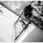 Black and white photo of a small dog looking down from a balcony. The balcony has a metal railing and is adorned with various hanging clothes. The sky is partly cloudy, and part of the building's wall is visible. The perspective is from below the balcony.