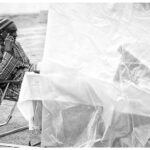 A black-and-white image of a seated person using a cell phone, partially obscured by a clear plastic sheet. The person wears a striped beanie and a plaid long-sleeved shirt and sits at a table covered with a cloth. The scene appears to be outdoors.