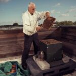 An older man wearing a beekeeping suit is standing on a rooftop holding a frame from a beehive filled with bees. He is inspecting the frame beside an open hive. Nearby, there are beekeeping tools and equipment, including a smoker and a tarp. The sky is partly cloudy.