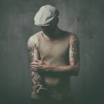 A person wearing a flat cap and a beige tank top looks down while adjusting their left sleeve. The person has intricate tattoos on both arms. The background is a textured, dark gray wall, creating a moody and dramatic atmosphere.