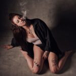 A person with long hair tied up is sitting on the floor in a relaxed pose, exhaling smoke. They are wearing a black shirt and light-colored underwear. They have multiple tattoos on their arms, legs, and chest. The background is a plain, dark gray wall.