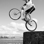 A black-and-white photo of a cyclist wearing a helmet and sports gear performing a wheelie on a mountain bike. The rider balances on the rear wheel atop a large concrete block, with an industrial cityscape and cloudy sky in the background.