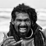 Black and white photo of a man with dreadlocks, smiling widely. He is making a shaka sign (thumb and pinky extended, other fingers curled) commonly associated with surfing culture. The background appears to show the ocean and blurred waves.