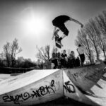 A skateboarder performs a trick in mid-air at a skate park under a bright sun. Onlookers sit and stand on the ramps, some watching intently. Bare trees and graffiti decorate the background, providing a dynamic urban atmosphere.