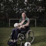 An elderly man sitting in a wheelchair on a grassy field holds a soccer ball on his lap. He is wearing a green and white scarf and there is an additional soccer ball near his wheelchair. Trees and a soccer goalpost are visible in the background.