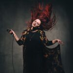 A person with long, vibrant red hair is captured in mid-motion, with hair fanned out dramatically. They wear glasses and a dark outfit featuring a patterned, flowing coat. They hold a microphone in their right hand and pose against a dark, simple background.