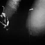 A person stands on a dimly lit stage holding an electric guitar, with a microphone stand in front. Smoke or mist surrounds them, creating a moody atmosphere. The lighting casts dramatic shadows, highlighting the person's silhouette.